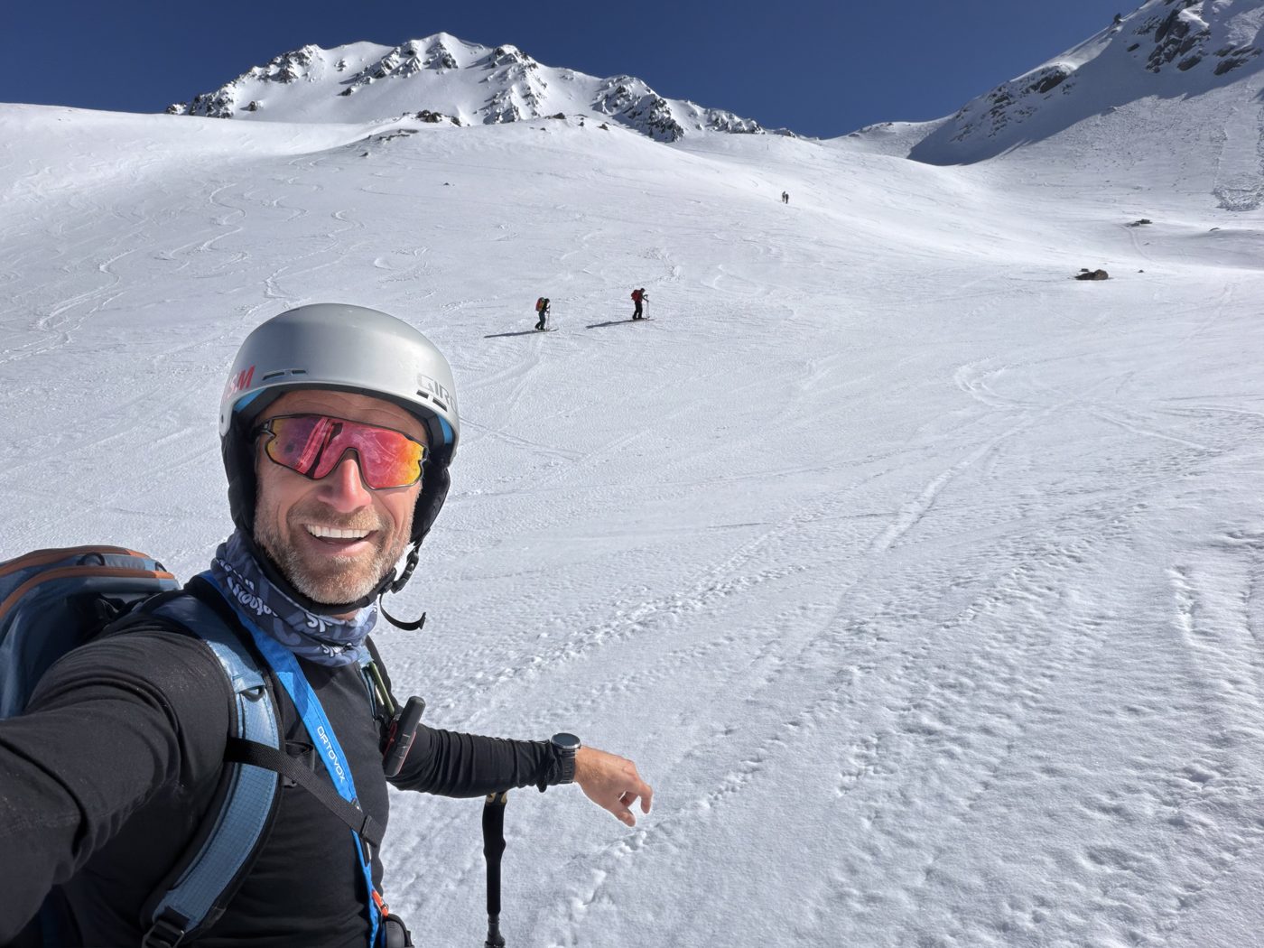 A ski tourer smiling on a sunny snow slope, with others skinning up towards the peaks behind